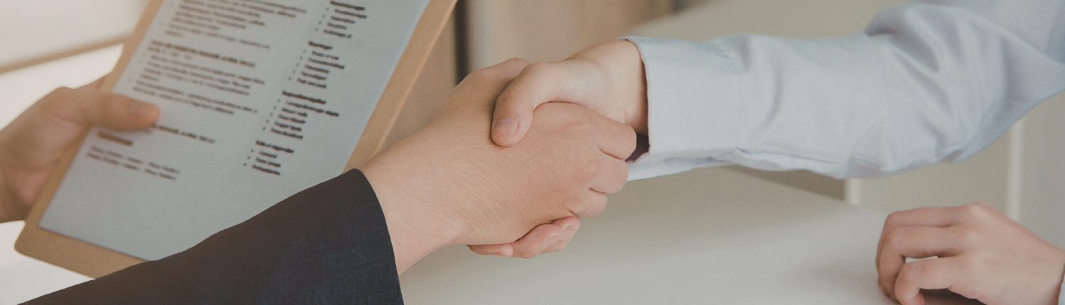 Two people shaking hands across a desk during an interview, with a CV visible in the foreground.