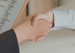 Two people shaking hands across a desk during an interview, with a CV visible in the foreground.