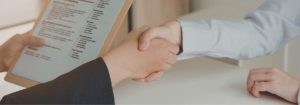 Two people shaking hands across a desk during an interview, with a CV visible in the foreground.