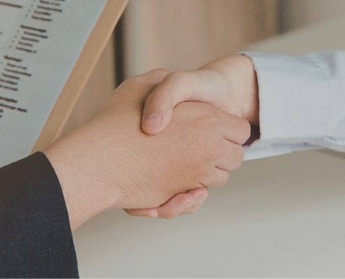 Two people shaking hands across a desk during an interview, with a CV visible in the foreground.