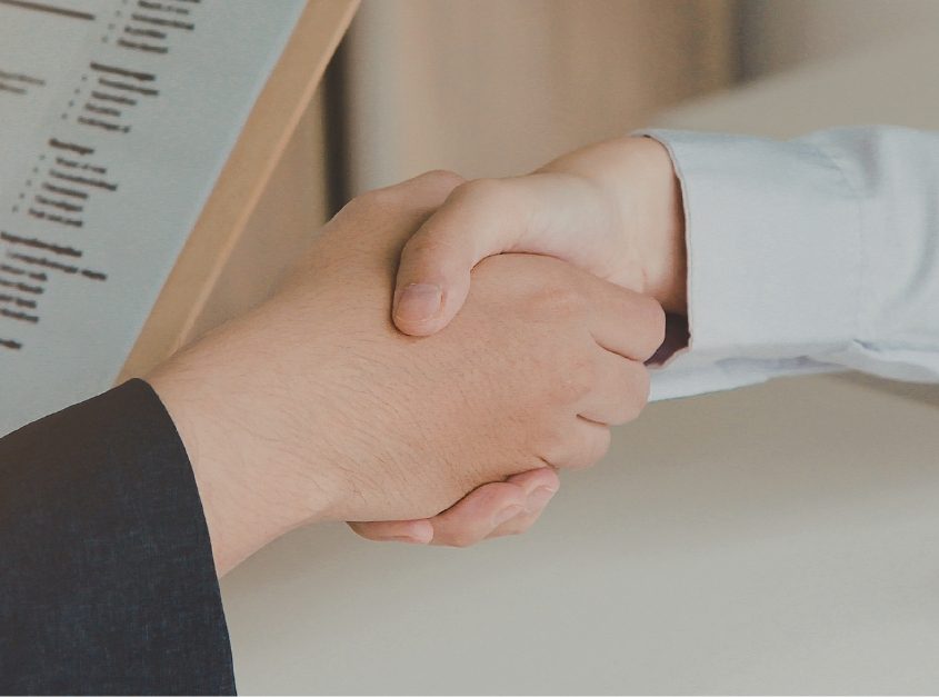 Two people shaking hands across a desk during an interview, with a CV visible in the foreground.