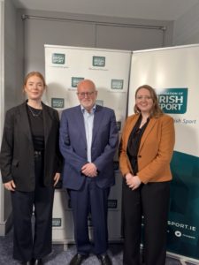Three people pose for a photo in front of Irish Sport banners; two women in black outfits flank a man in a blue suit.