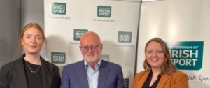 Three people pose for a photo in front of Federation of Irish Sport banners; two women flank a man in a blue suit.