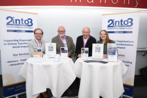 Four people stand behind a round table at a 2into3 booth, with banners saying 'Transforming Social Impact' and 'Since 2006'.