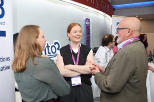A woman wearing a lanyard talks with two visitors at a booth during a conference.